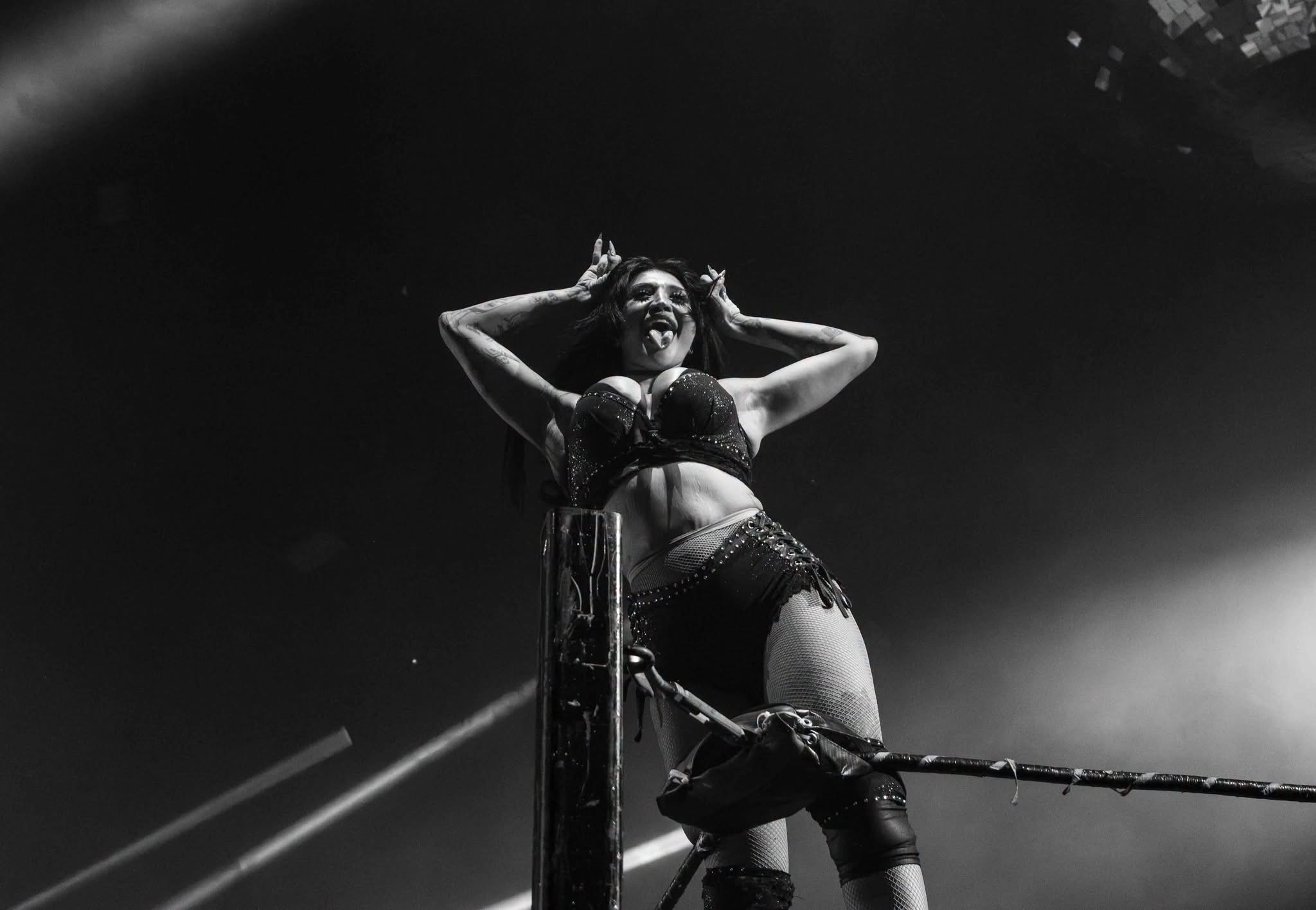 Black and white photo of Gypsy Mac standing on the turnbuckle making horn hand signs with her tongue out