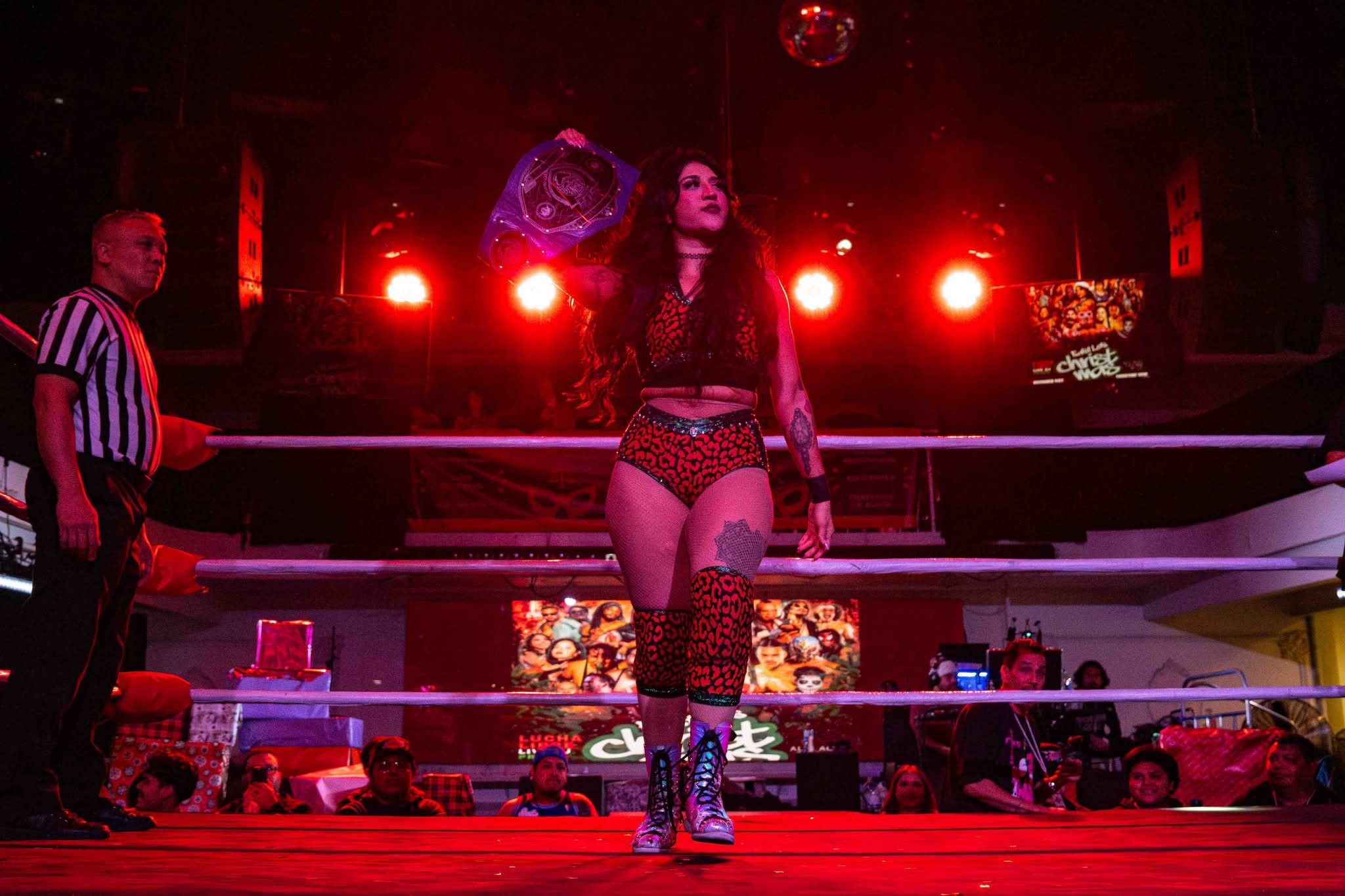 Gypsy Mac walking the ring in red light, holding a championship belt over her shoulder