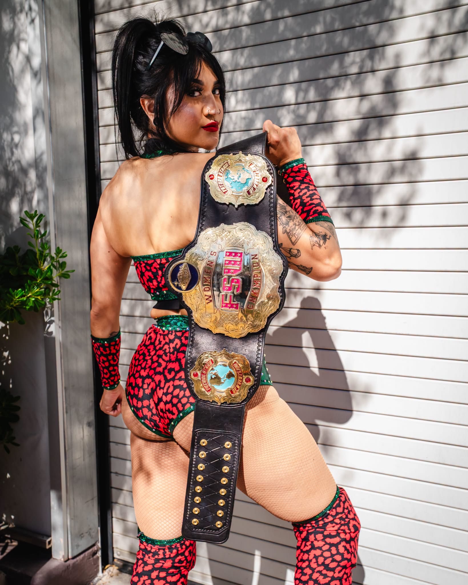 Gypsy Mac in red leopard-print gear looking over her shoulder, holding a gold championship belt against a white wall