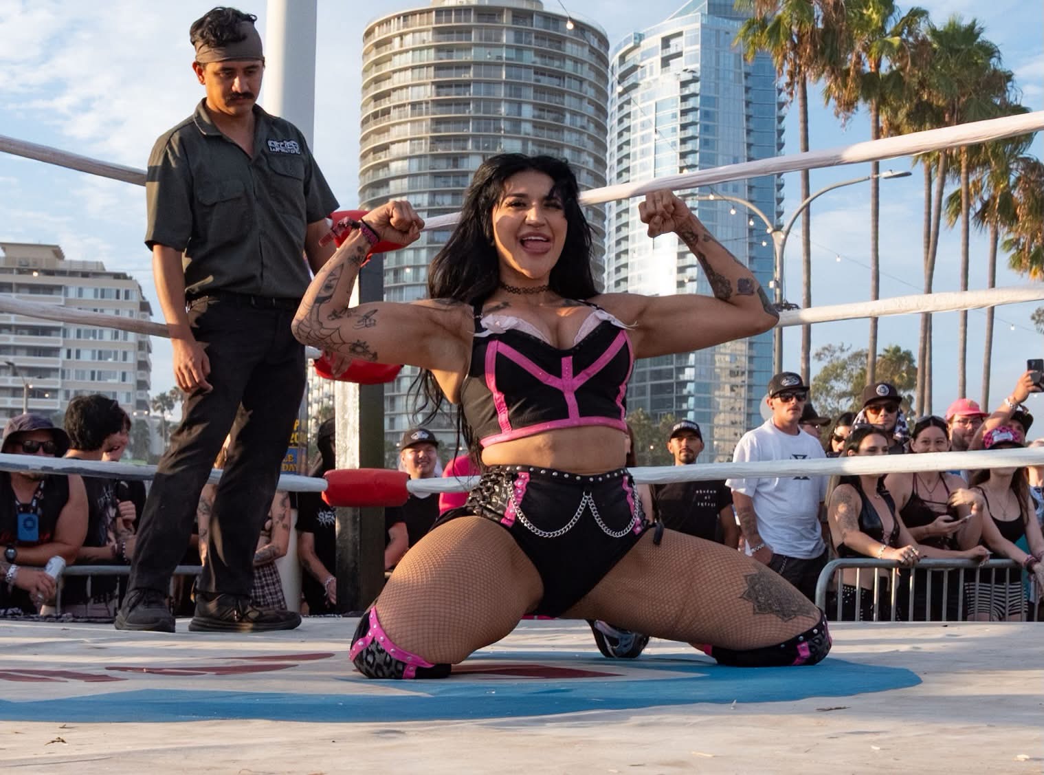 Gypsy Mac in black and pink gear kneeling in an outdoor ring and flexing for the crowd