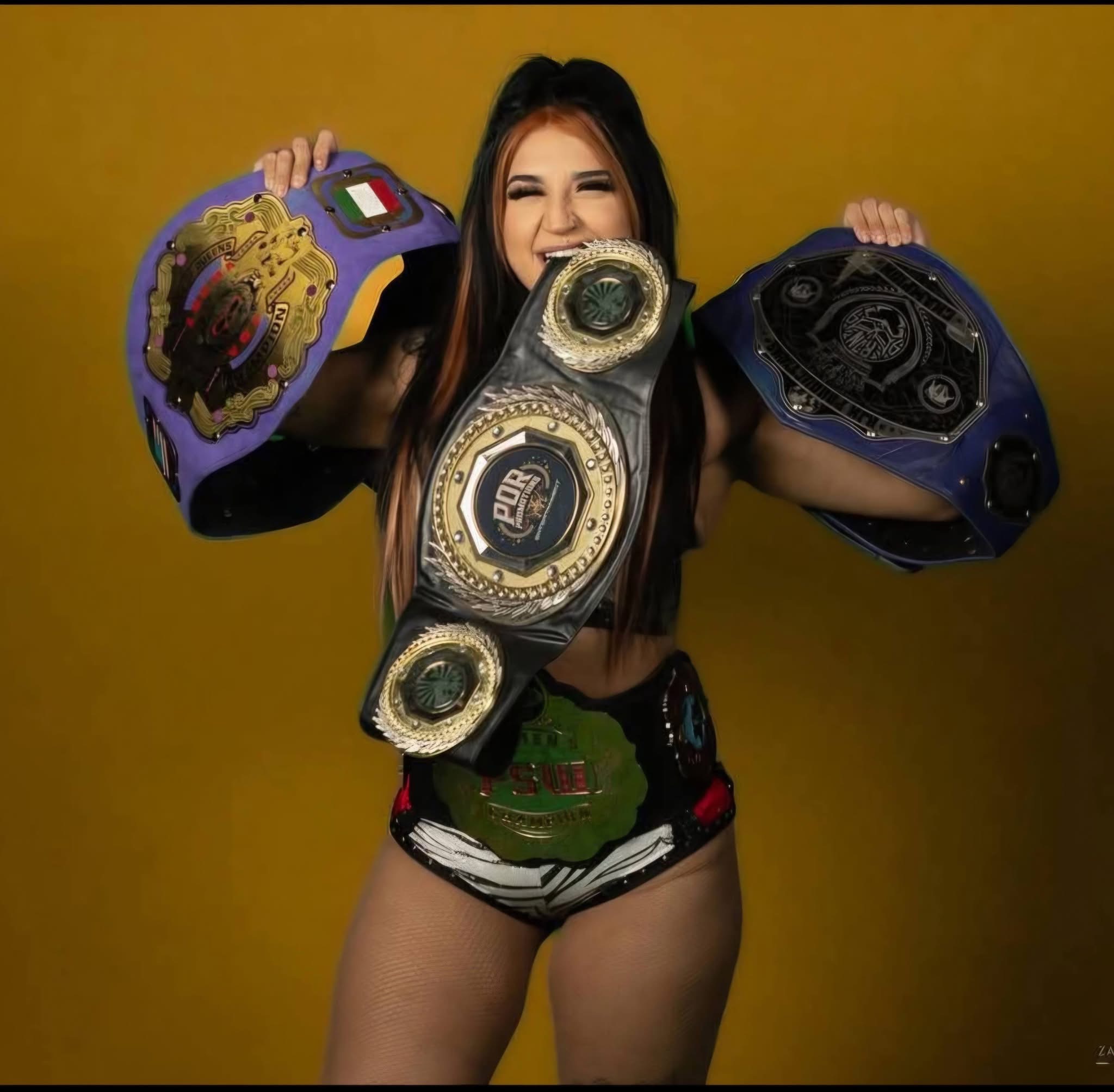 Gypsy Mac grinning against a gold backdrop, holding three championship belts on her shoulders and around her neck
