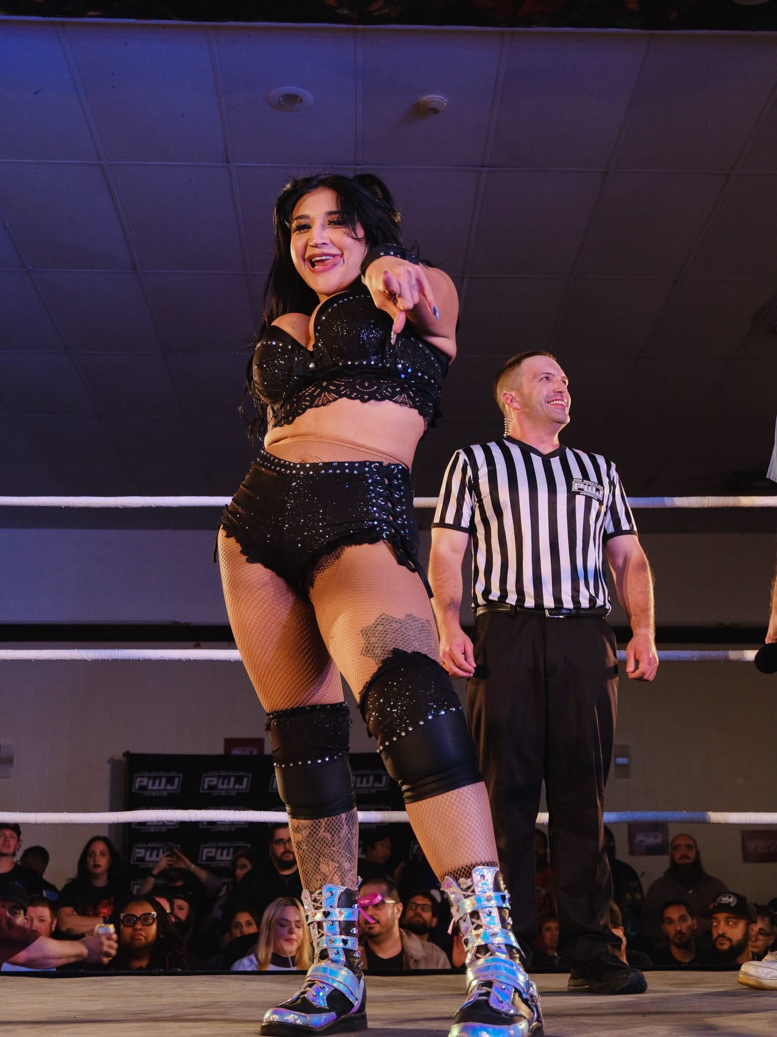 Gypsy Mac in black sparkly gear and chrome boots pointing at the camera in the ring, with a referee and crowd behind her