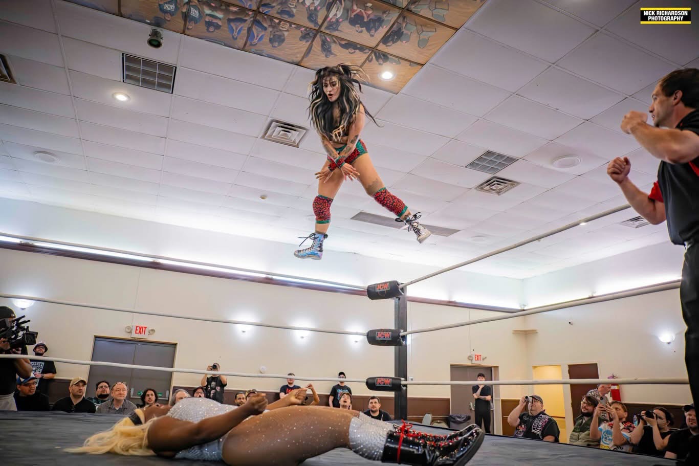 Gypsy Mac flying through the air toward an opponent lying on the mat, as the referee watches. Photo by Nick Richardson Photography