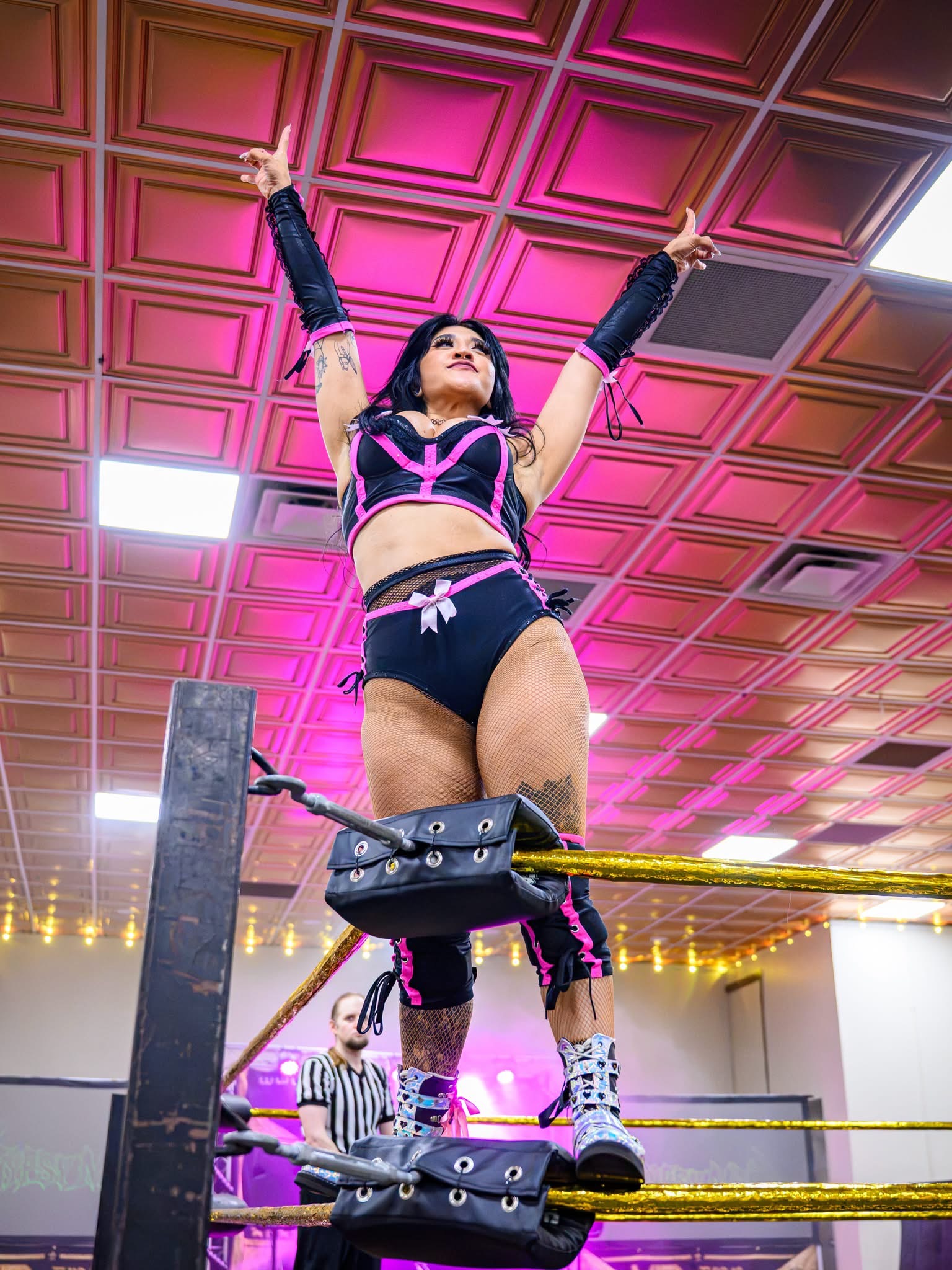 Gypsy Mac in black and pink gear standing on the top turnbuckle with both arms raised under a pink-lit ceiling